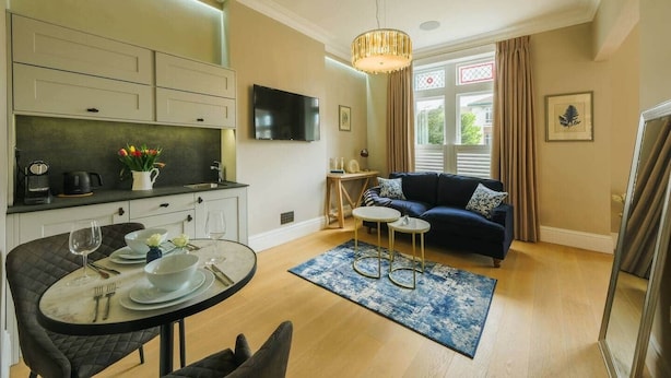 A modern living room with a dark blue sofa, a small round table, and a kitchen area with a TV above the counter. - Victorian Charm in Cotham | Free Parking (Bristol)