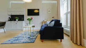 A modern living room with a dark blue sofa, a small round table with a vase of flowers, and a large window with beige curtains. - Victorian Charm in Cotham | Free Parking (Bristol)