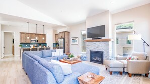 A spacious living room with a large blue sectional sofa, a fireplace, a television mounted above it, and a kitchen in the background. - Super clean 3-bedroom townhome- Purgatory - Hot Tub/Views/Arcade Game/Firepit (Durango)
