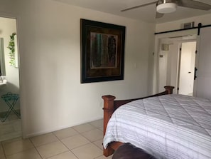 A bedroom with a bed, a ceiling fan, a framed artwork on the wall, and a door leading to another room.