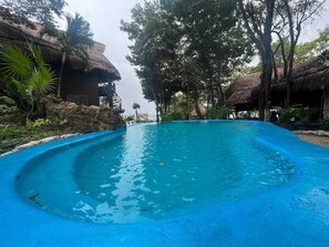Una piscina de borde curvo, rodeada de árboles y estructuras con techo de paja.