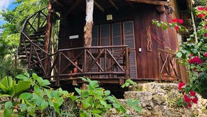 Una cabaña con techo de paja, balcón de madera y base de piedra, rodeada de exuberante follaje.