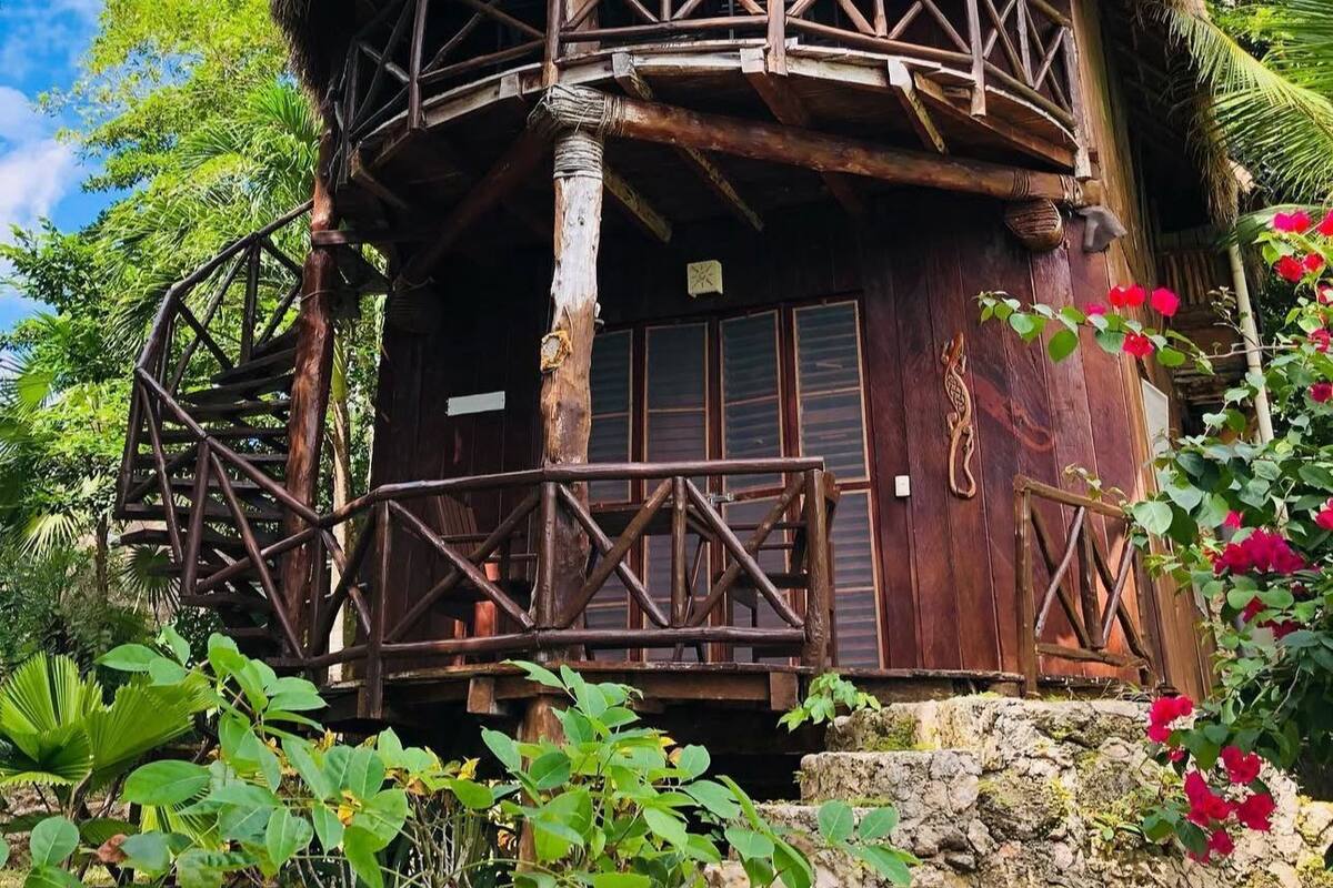 A thatched-roof hut with a wooden balcony and a stone base, surrounded by lush greenery.