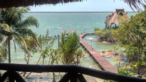 Un muelle de madera que conduce a una cabaña con techo de paja junto a un mar tranquilo, rodeada de exuberante vegetación.