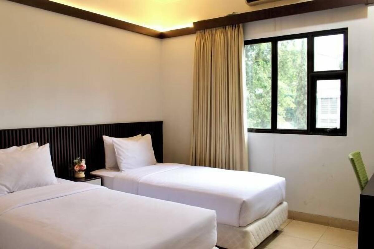 A hotel room with two beds, a nightstand, a window with curtains, and a view of greenery outside.