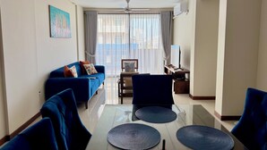 Een moderne woonkamer met een blauwe bank, een eettafel met stoelen, een plafondventilator en een groot raam met gordijnen.