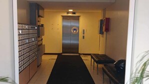 Un pasillo con alfombra negra que conduce a un ascensor. Hay armarios de almacenamiento a la izquierda y un banco a la derecha.
