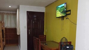 Un comedor con mesa y sillas de madera, un ventilador de pie y un televisor montado en la pared.