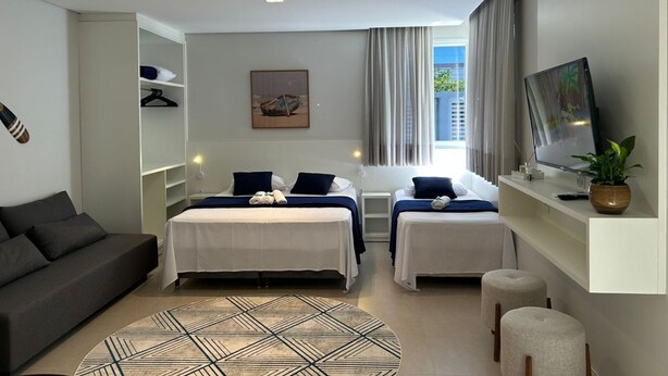 A modern hotel room with a bed, two bedside tables, a TV, a sofa, and a large window with curtains. - D'Santa Lofts - Penha - SC (Penha)