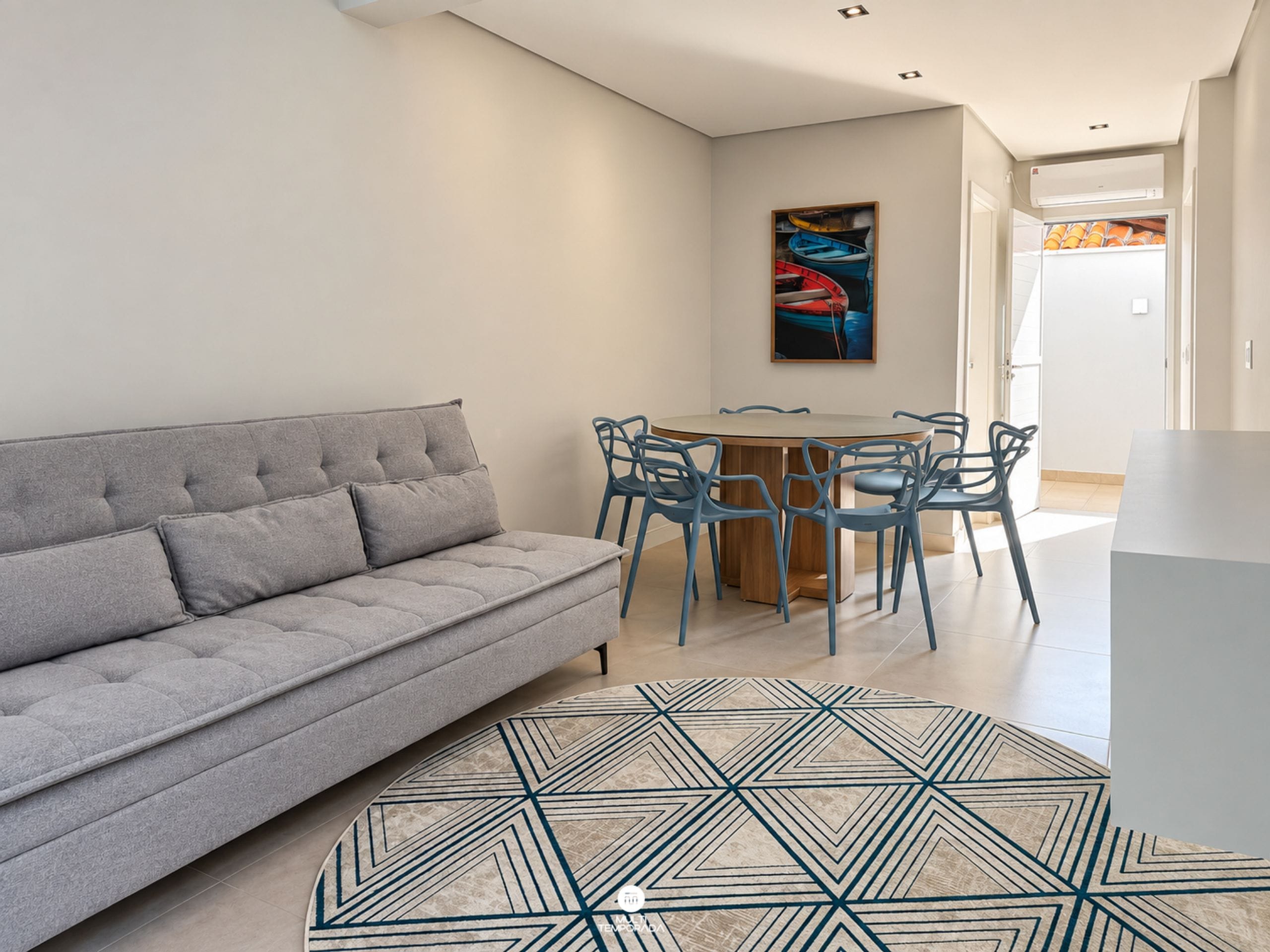 A modern living room with a grey sofa, a round wooden table with blue chairs, and a geometric patterned rug.
