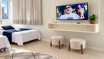 A modern hotel room with a bed, a TV mounted on the wall, a small seating area, and a view of the city through the window.