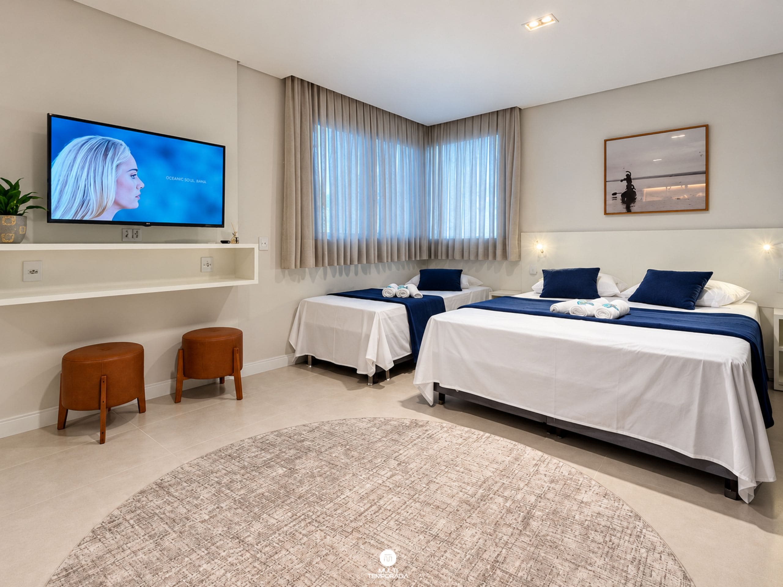 A hotel room with two beds, a TV, a large window with curtains, and a round rug.