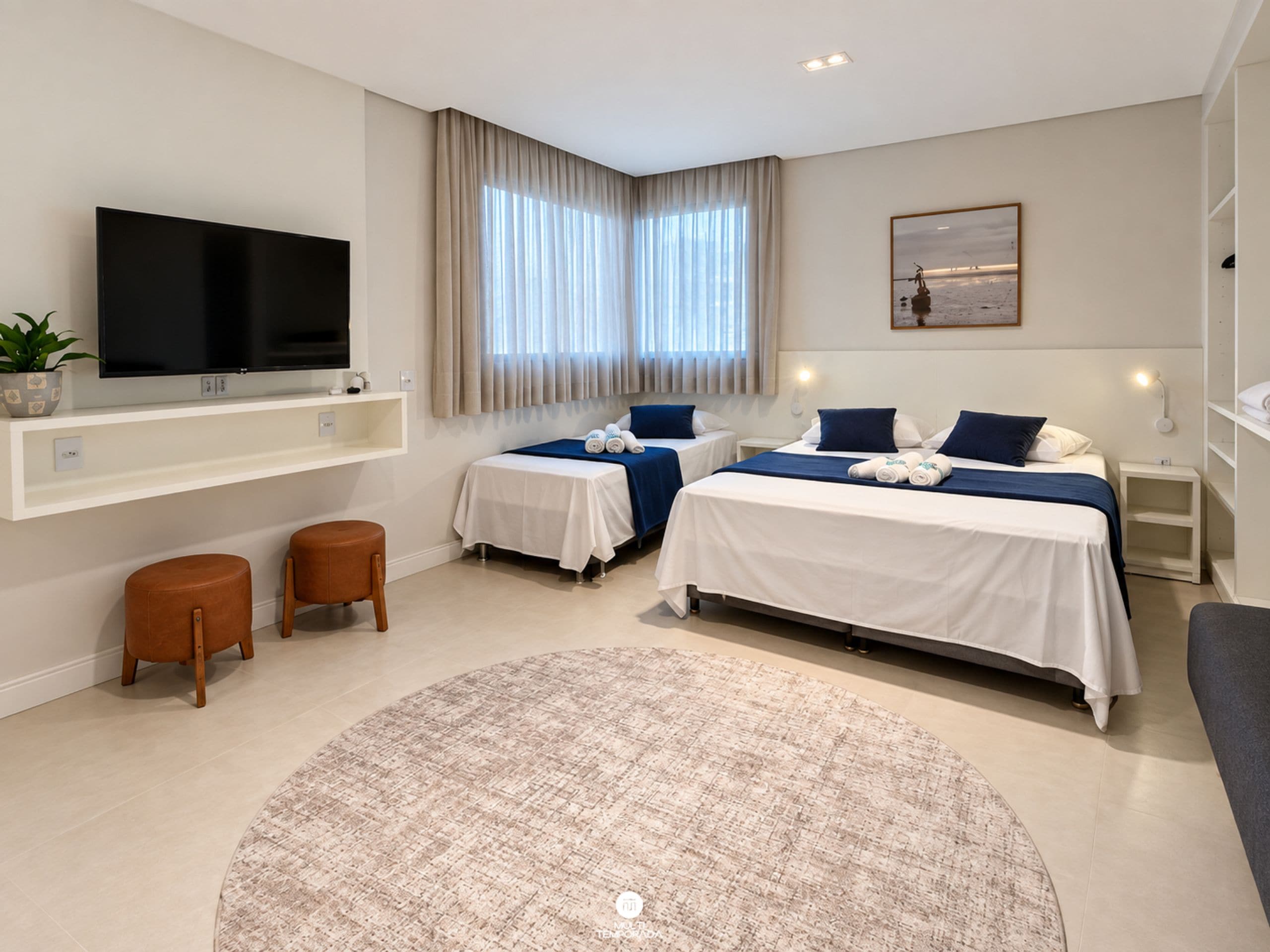 A modern hotel room with a flat-screen TV, two beds, a large window with curtains, a round rug, and a small plant.