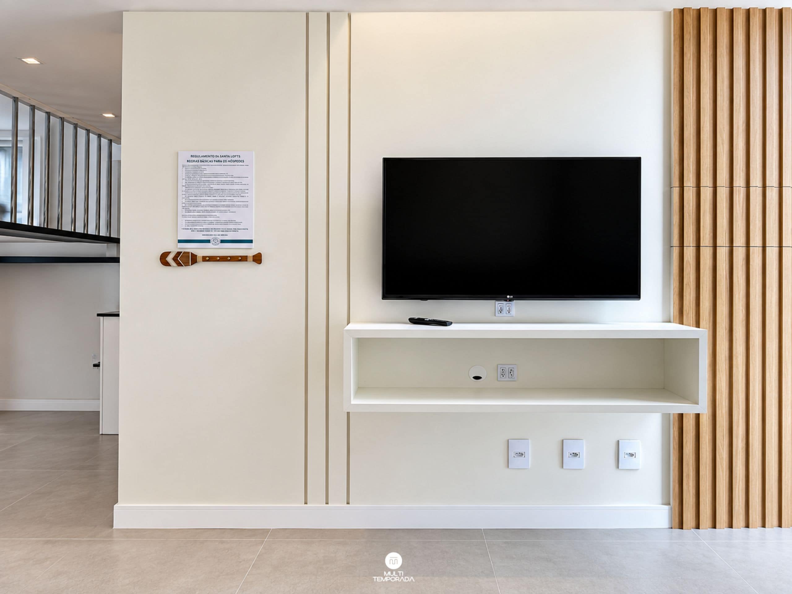 A modern living room with a flat-screen TV mounted on a white wall, a wooden slat wall panel to the right, and a small shelf below the TV.
