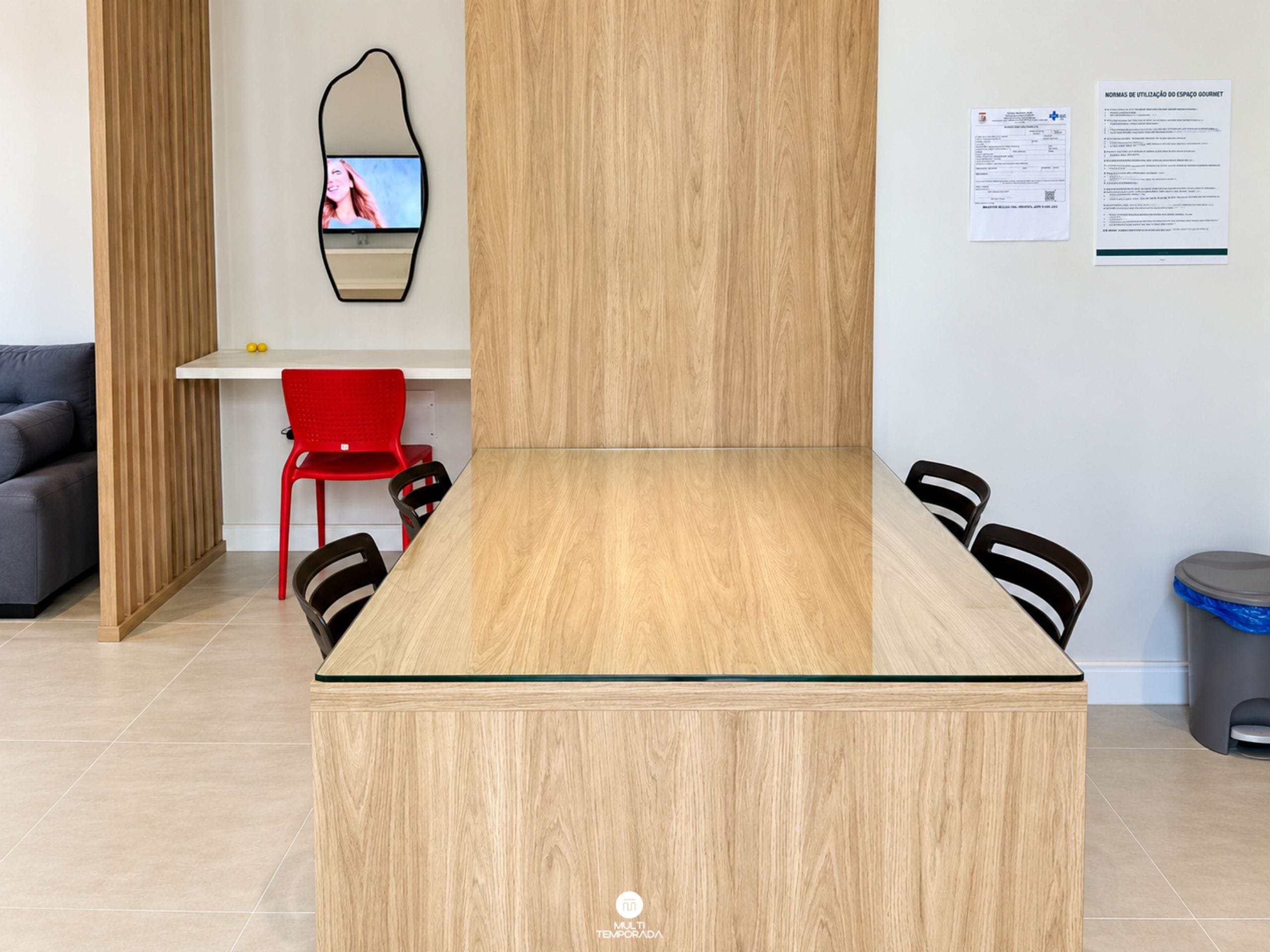 A modern meeting room with a wooden table, black chairs, a red chair, a white desk, and a mirror on the wall.