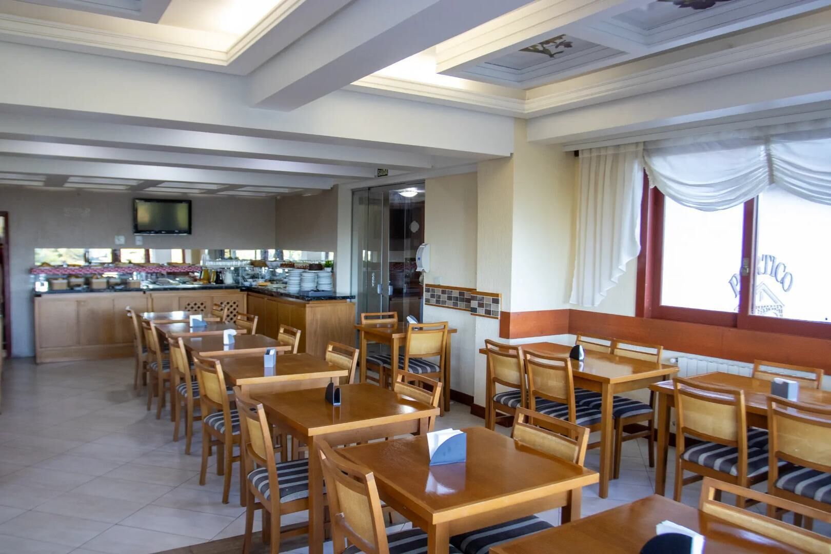 A restaurant with wooden tables and chairs, a kitchen area, and large windows with curtains.