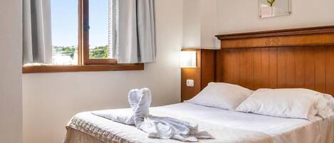 A hotel room with a bed, a window with curtains, a wall-mounted light, and a framed picture.