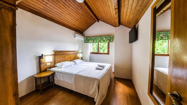 A bedroom with a wooden ceiling, a bed with a white bedspread, a bedside table, a window with a green valance, and a door. - HANNA Pórtico Hotel - Gramado (Gramado)