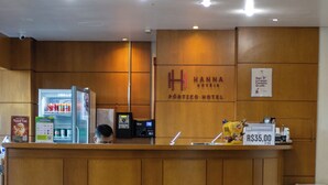 Un comptoir d’accueil avec une enseigne indiquant « Hanna Pórtico Hotel », une caisse enregistreuse, une poubelle et un réfrigérateur contenant des boissons.