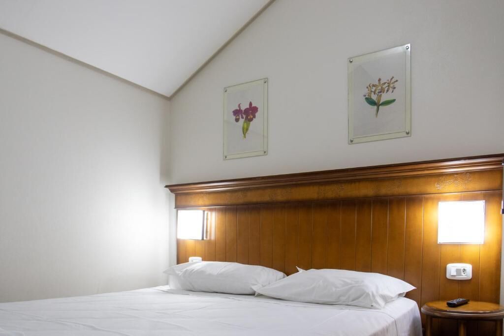 A hotel room with a wooden headboard, a bed with white bedding, two framed floral pictures on the wall, and a bedside table with a remote control.