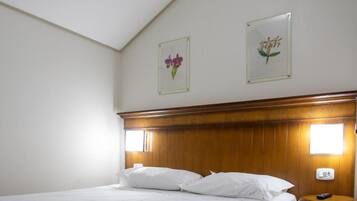 A hotel room with a wooden headboard, a bed with white bedding, two framed floral pictures on the wall, and a bedside table with a remote control.