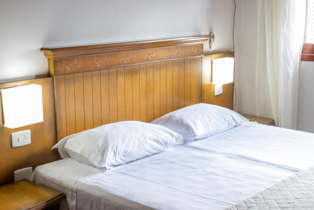 Habitación de hotel con cabecera de madera, dos almohadas blancas, una mesita de noche de madera y una colcha blanca.