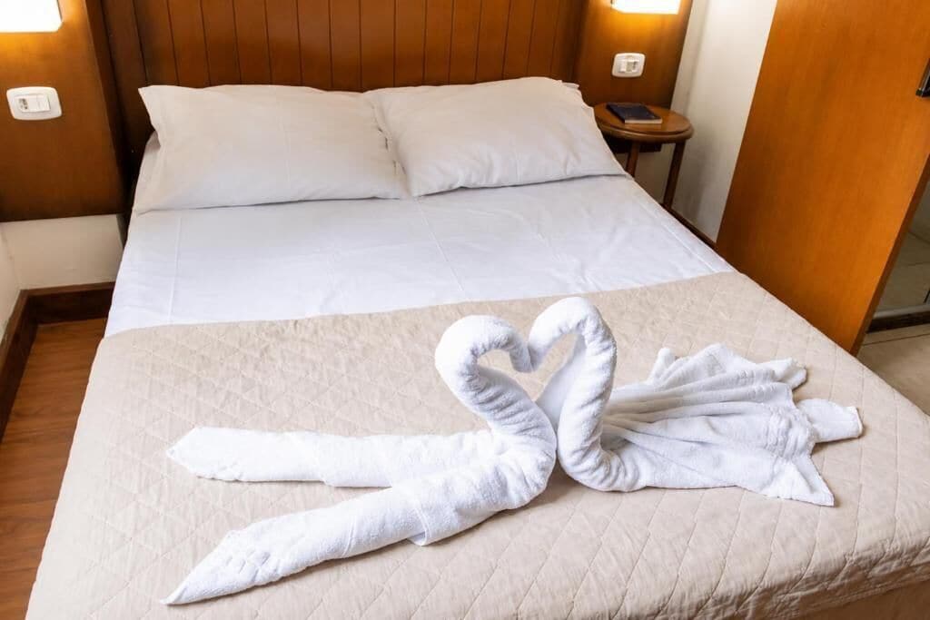 Quarto de hotel com uma cama, almofadas, mesa de cabeceira com telefone e luminária na parede.
