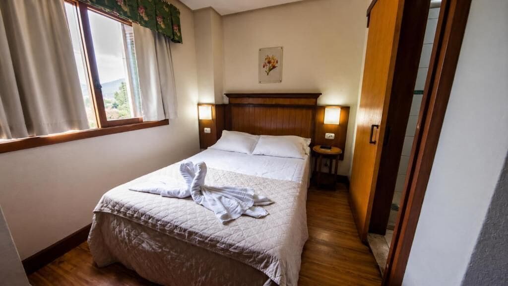 A hotel room with a bed, bedside lamps, a window with curtains, and a wooden floor.
