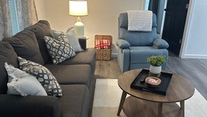 Un salón con un sofá marrón oscuro, un sillón azul, una mesa de centro redonda con un cuenco y un control remoto, y una lámpara sobre una mesita auxiliar.