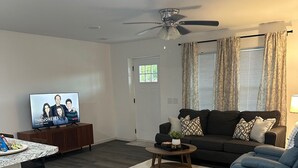 Un salón moderno con suelo de madera oscura, un sofá negro, un sillón azul, una mesa de centro y un televisor sobre un mueble de madera.