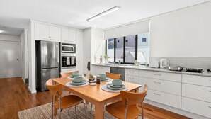 A modern kitchen with white cabinetry, stainless steel appliances, and a dining area with a wooden table and chairs. - Golden Views Hideaway - Beachside Apartment (Golden Beach)