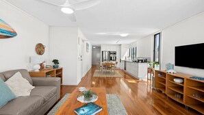 A modern living room with a sofa, coffee table, and TV. A kitchen with a dining area is visible in the background. - Golden Views Hideaway - Beachside Apartment (Golden Beach)