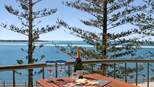 A table with a bottle of champagne, two glasses, and a plate of cheese and crackers, overlooking a beach with ocean waves. - Golden Views Hideaway - Beachside Apartment (Golden Beach)