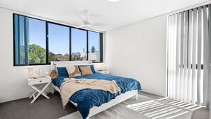 A modern bedroom with a bed, nightstands, a ceiling fan, large windows, and a view of the outdoors. - Golden Views Hideaway - Beachside Apartment (Golden Beach)