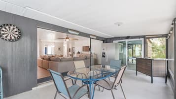 A modern living room with a glass dining table and chairs, a dartboard, and a view of the kitchen and dining area.
