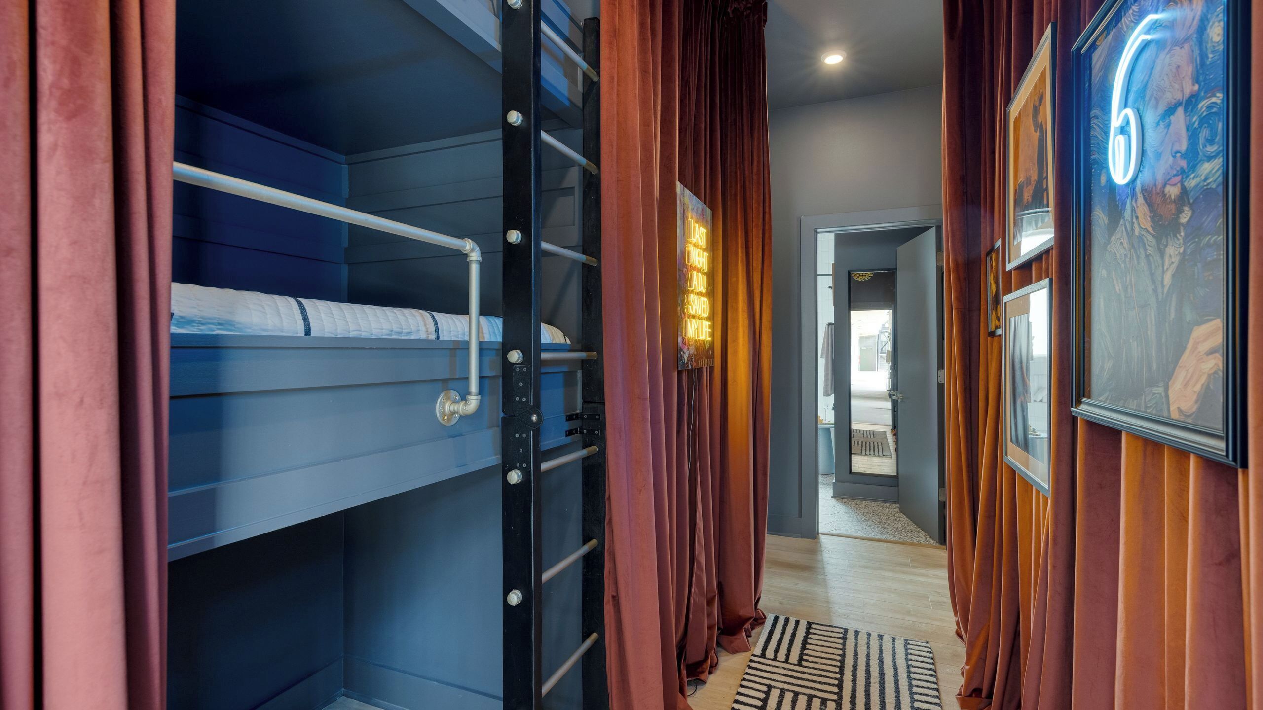 Une pièce avec un lit superposé, une porte ornée d’un néon, un plancher en bois et un couloir agrémenté d’un tapis à motifs.