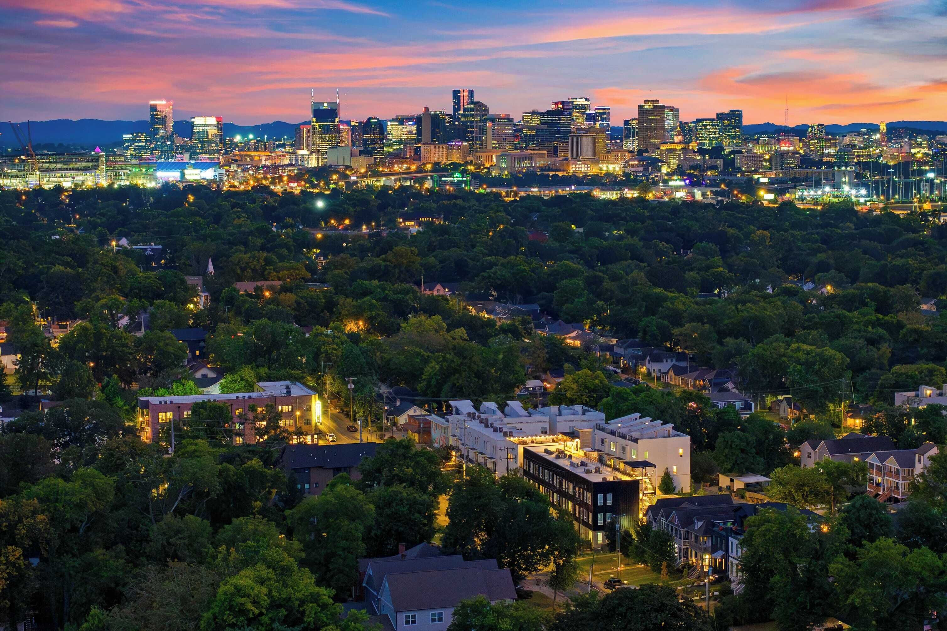 A cityscape at dusk with a skyline of illuminated buildings, a dense residential area below, and a river in the foreground.