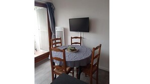 A dining area with a round table, four wooden chairs, a flat-screen TV mounted on the wall, and a window with curtains. - Apartment on the heights of Gérardmer with a superb view (Gérardmer)