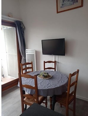 A dining area with a round table, four wooden chairs, a flat-screen TV mounted on the wall, and a window with curtains. - Apartment on the heights of Gérardmer with a superb view (Gérardmer)