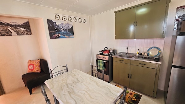 Cocina compacta con estufa, lavamanos y armarios. Se observa una cama con almohada y un cuadro colgado en la pared.