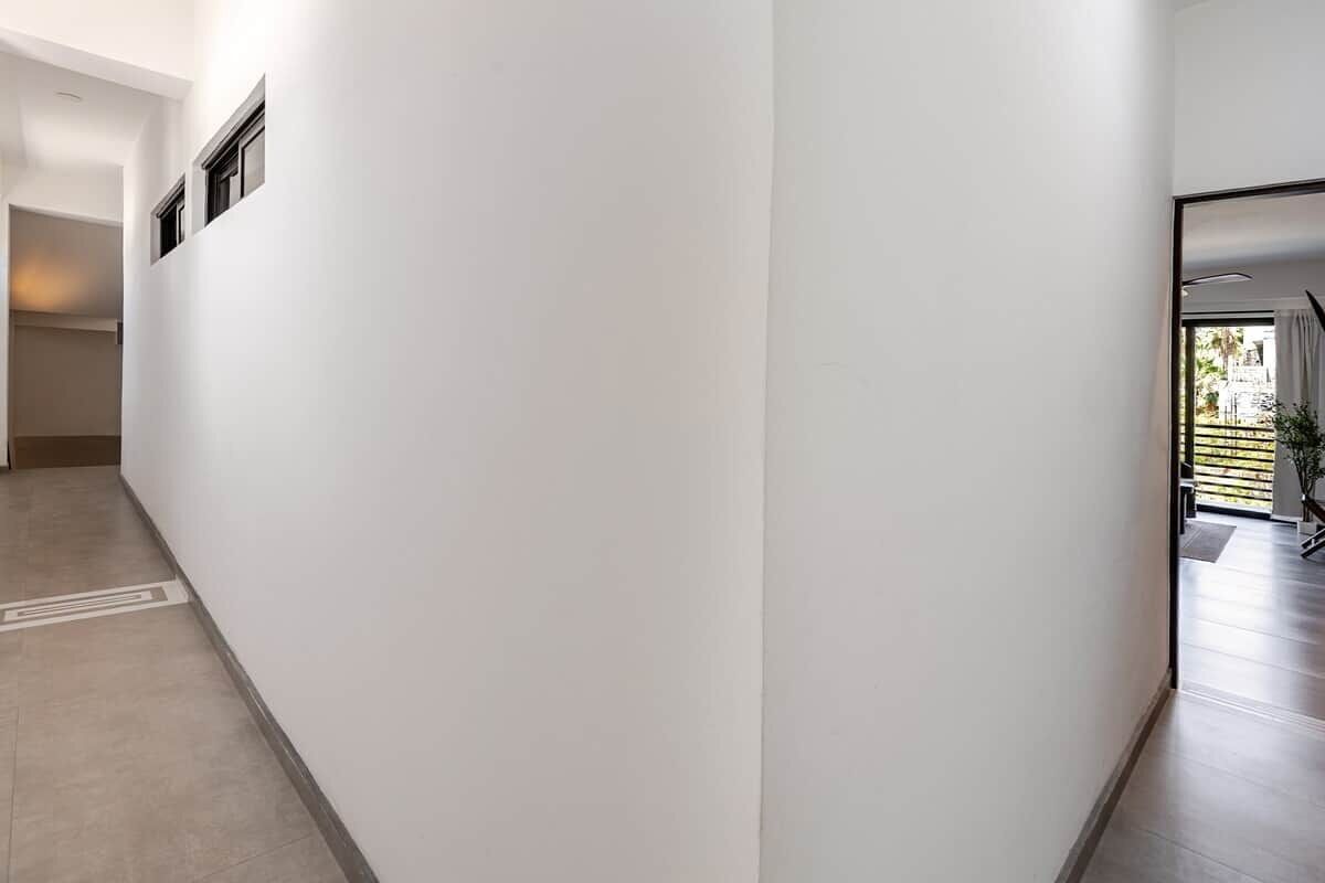 A hallway with white walls and a tiled floor, leading to a room with a large window and a plant.