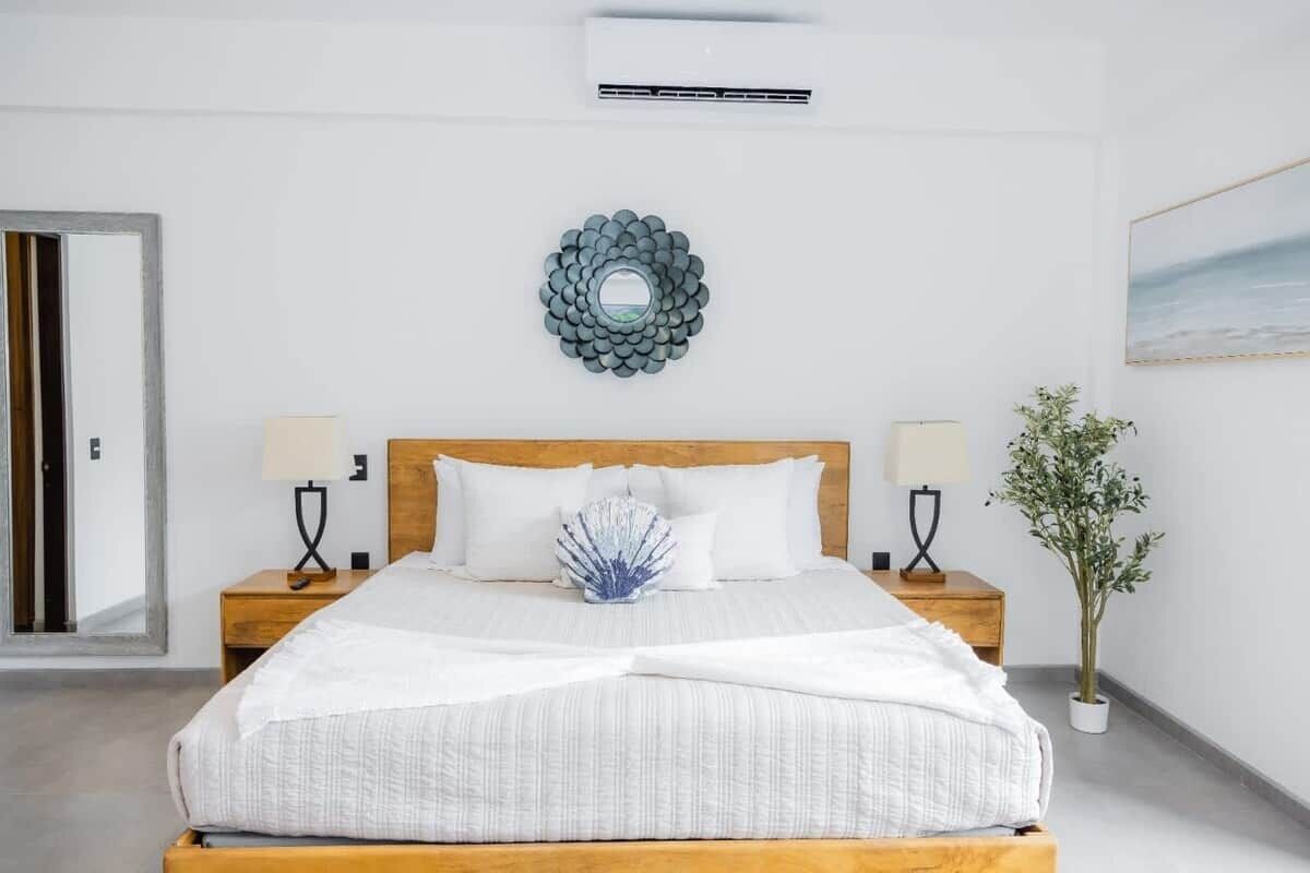 A hotel room with a bed, two bedside tables, a wall-mounted air conditioner, a decorative wall piece, and a potted plant.