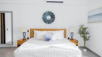 A hotel room with a bed, two bedside tables, a wall-mounted air conditioner, a decorative wall piece, and a potted plant.