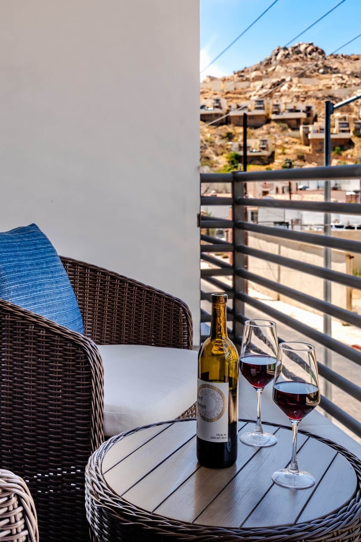 A wicker chair with a cushion, a bottle of wine, and two wine glasses on a balcony table.