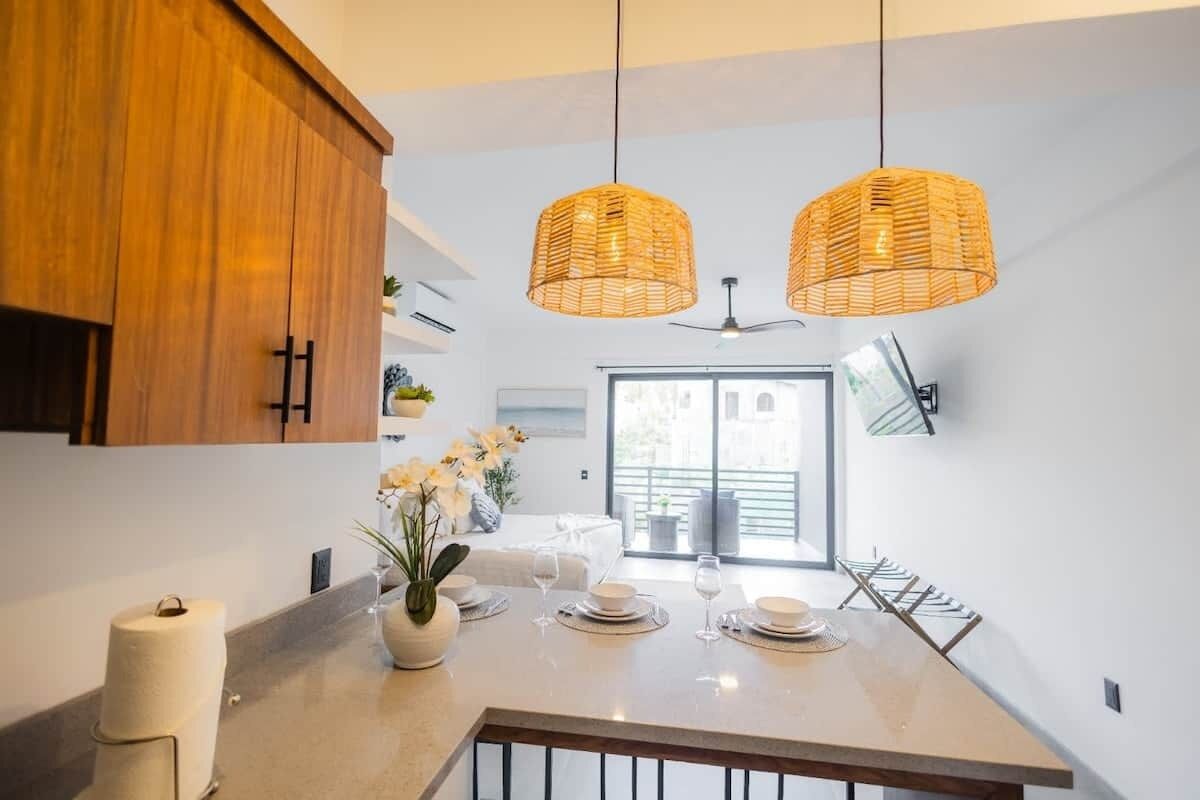 A modern kitchen with wooden cabinets, a central island with a vase of flowers, and two hanging wicker pendant lights.