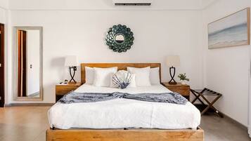 A bedroom with a wooden bed, two bedside tables, a wall-mounted air conditioner, a decorative mirror, and a painting on the wall.