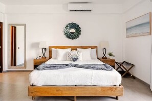 A bedroom with a wooden bed, two bedside tables, a wall-mounted air conditioner, a decorative mirror, and a painting on the wall.