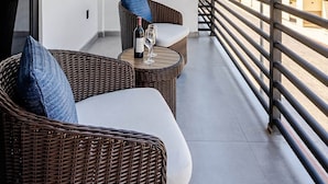 Un balcon agrémenté de meubles en osier, une table avec une bouteille et des verres, et une vue sur des bâtiments au loin.