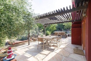 Un patio exterior con pérgola, suelo de piedra, mobiliario de madera y zona de barbacoa.