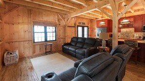 Interior acogedor de una cabaña de madera con sala de estar que incluye sofás de cuero, comedor con cocina y un gran ventanal que proporciona luz natural.